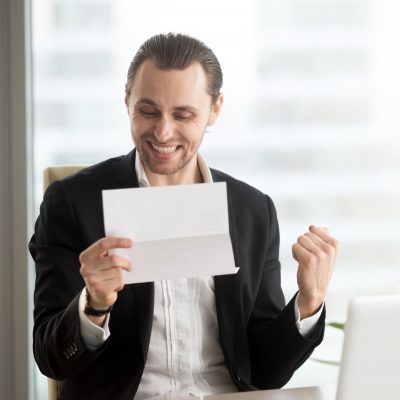 Happy smiling businessman celebrates receiving good business news in letter while sitting in front of laptop in modern office. Successful deal, bonus or salary raise, financial offer accepted concept.