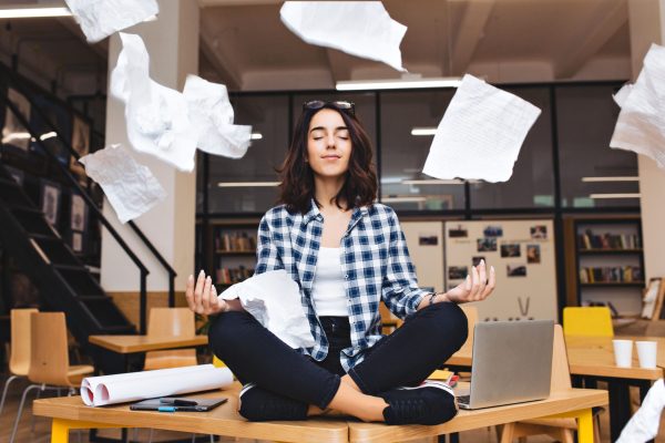 Young pretty joyful brunette woman meditating on table surround work stuff and flying papers. Cheerful mood, taking a break, working, studying, relaxation, true emotions